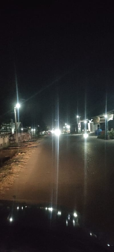A nighttime street scene in Blangcot Tunong, Indonesia. The image appears to have been taken from a vehicle looking down a road. Streetlights illuminate the roadway, with some buildings and foliage visible on either side. Headlights from distant vehicles are also visible. The foreground is dark, likely the dashboard of the vehicle.