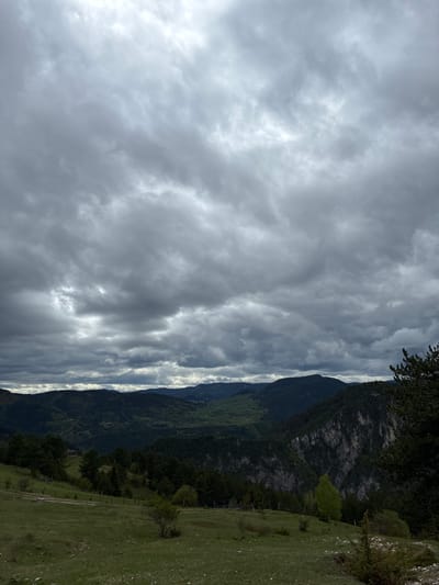 The image shows a landscape in Giovren, Bulgaria under a heavily clouded sky. Mountains covered in green vegetation rise along the right side of the frame. A grassy field extends to the lower right. The sky takes up a significant portion of the image with thick, gray clouds filling the space.