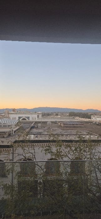 The image, when properly oriented to a landscape view, presents a wide panoramic scene at either sunrise or sunset. On the far left, a dark, textured wall or pillar of an unseen structure occupies the foreground. To its right, the sky transitions from a soft light blue at the top to warm shades of orange and yellow closer to the horizon, indicating the early morning or late evening.

In the midground and background, a sprawling landscape unfolds. Distant, undulating hills or mountains form a subtle ridge line against the colorful sky. Below these natural contours, an urban or developing area is visible. Several white buildings with distinctive architecture, including prominent arched windows and decorative patterns, are scattered across the scene. One building, particularly noticeable, features a large, ornate archway. Adjacent to these structures, a significant portion of the land appears cleared and flat, resembling a large construction site or undeveloped plot of earth, with areas of exposed soil.

On the far right side of the image, a large, reflective glass surface, likely a window or a building's facade, dominates. This surface strongly reflects numerous bare tree branches and some green foliage, suggesting trees are in close proximity. These reflections are so vivid that they largely obscure any view through the glass itself.

The scene depicts a quiet, expansive vista of a landscape that includes both natural elements and developing urban infrastructure. No specific human activity or visible text is present. The notable light of the sky suggests a peaceful moment at the beginning or end of the day. The location context provided is Tixeraïne, Algeria.