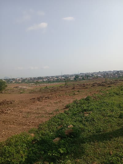 This image captures a panoramic view of a developing area on the outskirts of Tasha, Nigeria. The foreground is dominated by dry, brown earth, indicative of cleared land or agricultural fields. Scattered stones and some sparse vegetation are visible on this dry ground. Moving towards the midground, the terrain transitions to areas with more vibrant green, low-lying plants and grasses, suggesting either natural growth or a more cultivated patch.
Further into the distance, a sprawling settlement is nestled against a backdrop of gently rolling hills. The settlement consists of numerous residential buildings with varied roof colors, predominantly red and white, interspersed with trees. Several communication towers are also discernible within the cityscape, indicating the presence of infrastructure. The sky above is a hazy, pale blue with a few wispy clouds, suggesting a bright but possibly dusty atmosphere. The overall impression is one of rural expansion and the ongoing development of a residential area in a semi-arid or savanna environment.