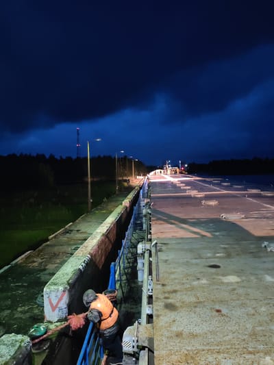 The image depicts a nighttime scene on a concrete structure, likely a bridge or a dock, in Devyatiny, Russia. The sky is a deep, dark blue, with heavy clouds obscuring most of the stars. A single person, wearing an orange life vest and a helmet, is actively engaged in pulling a thick, red rope. The rope appears to be secured to a concrete bollard marked with a red "V" symbol. The person is leaning over a blue metal railing, indicating they are working near the edge of the structure.

The concrete surface is worn and textured, with visible patches and stains. Several round hatches with metal clasps are embedded in the surface, suggesting a large, flat-topped vessel or platform. The scene is illuminated by artificial lighting, casting long shadows across the concrete. In the distance, there are lights indicating structures or vehicles, and a tall tower with red lights is visible on the horizon, silhouetted against the dark treeline. The environment appears to be a waterway, with dark water visible to the left of the concrete structure, which is bordered by a grassy area. The overall impression is one of an industrial or operational setting at night, with a focus on the activity of the lone worker.