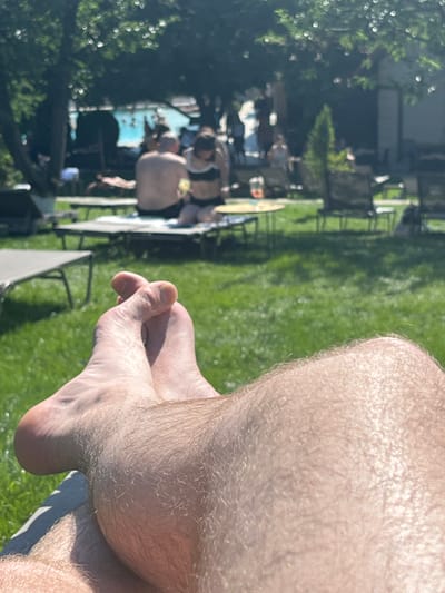 In Stăuceni, Moldova, a first-person perspective shot shows a person's bare feet crossed in the foreground, resting on green grass. Sunlight illuminates the hairy skin on their legs and feet. In the blurred background, several people are visible near a swimming pool. Some are sitting on lounge chairs, while others appear to be in or around the pool. The overall setting suggests a sunny day at an outdoor recreational area, possibly a resort or a private residence with a pool.