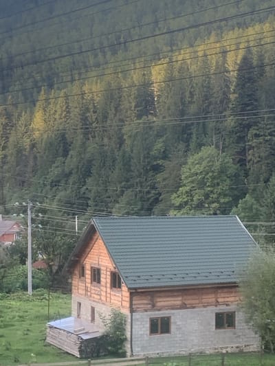 The image displays a two-story residential structure situated on a grassy, uneven slope in what appears to be Tatariv, Ukraine. The primary building features a dark green, corrugated metal roof with visible snow guards. Its upper level is constructed from horizontal light-colored wooden logs or planks, featuring multiple dark-framed windows, with three visible on the main facade and one on the gable end. The lower level is constructed from grey concrete blocks, with four dark-framed windows visible on the main facade. To the immediate left of the house, a stack of light-colored lumber is observed, partially covered by a transparent tarp. A low, wooden fence is partially visible in the lower right foreground.
In the midground, multiple utility poles are present, with numerous black electrical or communication wires extending horizontally across the frame. Portions of other residential buildings, including one with an orange-red roof, are visible to the left of the main structure. The background consists of a dense, steep hillside entirely covered by a mature coniferous forest, with areas of lighter green foliage indicating sunlight exposure. No individuals, animals, actions, or interactions are present in the scene.