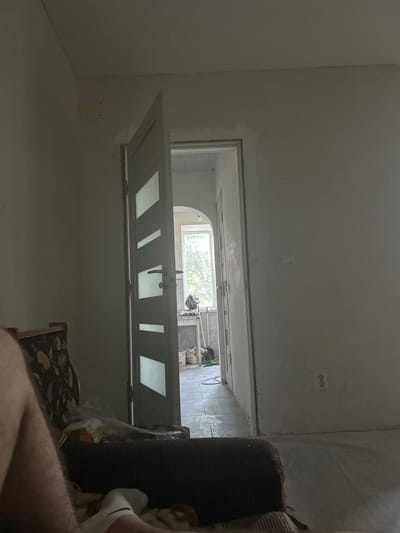 In a dimly lit room that appears to be undergoing renovation, a modern interior door with horizontal glass panels stands ajar, revealing a hallway or another room beyond. The walls are unfinished, with visible signs of plaster work. In the foreground, the corner of a dark upholstered armchair with a floral pattern is visible, and a person's legs and feet are also in view, suggesting someone is sitting or lying on furniture within the room. To the right, a white electrical outlet with two circular receptacles is mounted on the wall. Through the open doorway, a glimpse of a window with trees outside can be seen, as well as a table with some items on it, including buckets and cleaning supplies, indicating construction or cleaning activities. The floor inside the doorway appears to be tiled. The overall impression is of an interior residential space in Florești, Moldova, in a state of transition.