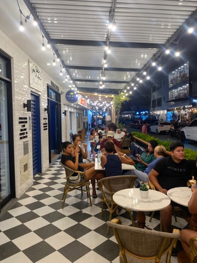 The image shows an outdoor dining area in Punta Hermosa, Peru (latitude: -12.333, longitude: -76.826). It is located on a street lined with businesses, with a canopy overhead that is lit with string lights. The dining area is furnished with tables and chairs. Several people are sitting at the tables, eating and drinking. The floor is tiled in a black and white checkerboard pattern. The street is paved with asphalt and there are cars parked on the side of the street. There are green plants along the edges of the dining area. The sky is dark, with a few stars visible.