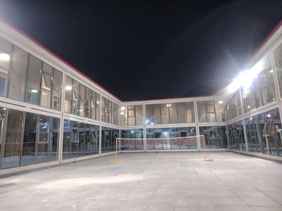 This media file depicts an outdoor badminton court at night, surrounded by a modern building with extensive glass facades. The building appears to be modular, with a white structure and red trim along the roofline. Many of the glass panels are open, suggesting ventilation or access points. Interior lights are visible through the windows, illuminating the space within.
The badminton net is set up in the center of the court, with yellow net posts. The court itself is paved with large, square tiles. The sky above is dark, indicating nighttime, and there are no visible weather conditions other than the clear darkness.
A single person is visible through the glass in one of the building sections, appearing to be standing or walking inside. The overall impression is one of a recreational facility or communal space attached to living quarters, perhaps associated with a dormitory or a workers' camp, given the functional design of the building. The bright interior lights contrast with the dark sky and the illuminated area of the court. There is no visible text in the image. The location context is Farab, Tajikistan.