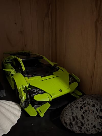 An unbranded, bright green, Lego Technic style toy car is displayed on a dark shelf, which is set against a wood-paneled wall. To the left of the toy car is a white, textured object resembling a large seashell. To the right of the car is a dark gray, porous rock. The lighting in the scene is dim and appears to be coming from the front and slightly above. The image does not contain any information about Sankt Augustin, Germany.