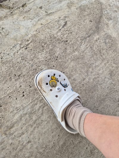 A close-up, overhead shot shows a child's foot wearing a white Croc shoe with a light brown sock. The Croc has two charms attached: one a cartoonish yellow duck-like figure, and the other a white button with the letter "m" in blue. The shoe appears slightly dirty. The foot and shoe are resting on a textured, grey concrete surface. The concrete has areas of lighter and darker patches, with small pits and rough edges, suggesting an outdoor environment. The lighting indicates it is daytime, with natural light casting subtle shadows, though the exact time of day is not discernible. The image's focus is on the shoe and the immediate surroundings, with the background gently blurred. There is no clear indication of a specific event or activity beyond the presentation of the shoe and its setting. The scene is neutral and lacks any discernible emotional tone. The context "Lenina, Russia" provides a geographical location but does not offer visual cues within the image itself to confirm this.