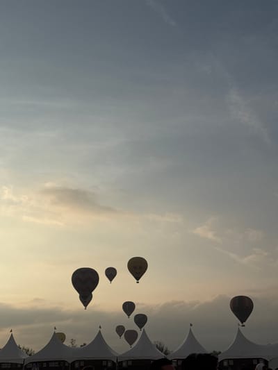 The image shows a sky scene with several hot air balloons floating in the air. The sky transitions from a light blue to a warmer, golden hue, likely indicating either sunrise or sunset. The hot air balloons are of varying sizes and designs. On the right side of the image, there are partial views of white tents or canopies with people standing underneath or near them. The location is suspected to be San Francisco Mazapa, Mexico, but the image itself does not provide definitive proof of this.