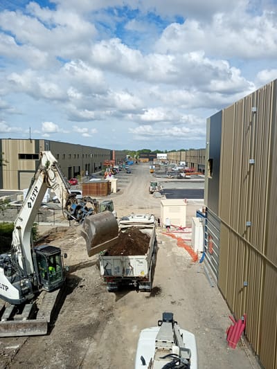 The image shows a construction site in Mérignac, France, with several large industrial buildings under construction or recently completed. In the foreground, a white excavator with the "LTP" logo is actively digging and loading dirt into a white Volvo dump truck. The excavator's bucket is full of dark soil, and some soil has spilled onto the ground. A worker in a high-visibility vest is visible in the excavator's cab. The Volvo truck has French license plates and the "TML" logo on its side.
The construction site is dusty and appears to be a work in progress. Further in the background, more industrial buildings are visible, stretching into the distance. Various other construction vehicles, including smaller excavators, a forklift, and a tractor, are scattered across the site. A line of parked cars and a few other trucks are visible along a paved road. The sky is partly cloudy with dramatic white cumulus clouds against a blue background. Orange construction netting is also visible in some areas. The architecture of the buildings suggests modern industrial or commercial facilities.