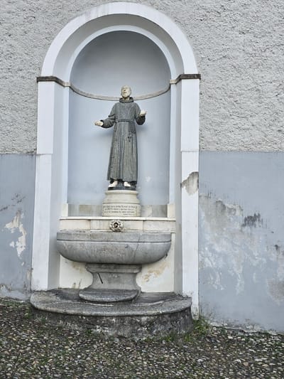 This media file features a stone statue of a robed figure, likely a saint or religious person, displayed within an arched niche. The statue, carved from light-colored stone, depicts the figure with outstretched arms and is adorned with a rosary around its neck. It stands in front of a cylindrical pedestal that bears inscribed text. The niche itself is framed by a white architectural element against a textured gray wall. To the right of the niche, a large, weathered stone urn or fountain base is visible, juxtaposed with a gravel pathway dotted with small stones and patches of grass. The overall scene suggests an outdoor religious or memorial setting. The lighting indicates daytime, with no specific weather conditions apparent. The visible text on the pedestal is in Italian and appears to be an inscription relating to the figure or the monument. The style of the statue and the architectural elements suggest a European location, consistent with the provided location context of Orselina, Switzerland.