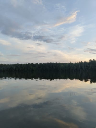 The image depicts a serene lake with a dense forest lining its far shore under a pale, cloudy sky. The water's surface acts as a mirror, reflecting the sky and the trees, creating a symmetrical composition. The scene appears tranquil and natural, with no people or specific man-made objects visible. The location is identified as Kichier, Russia, suggesting a natural landscape within that region. The time of day appears to be either dawn or dusk, indicated by the soft lighting and the muted colors in the sky and its reflection.