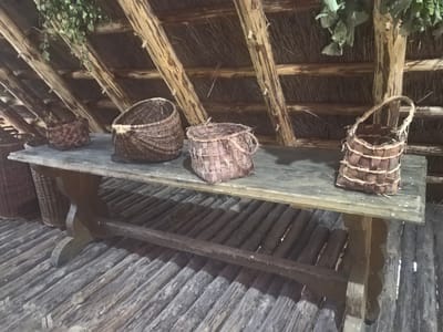 The image depicts a rustic interior scene, likely within a traditional dwelling or exhibit. A sturdy, dark wooden table with carved legs rests on a floor constructed from rough-hewn logs. On the table are several woven baskets of varying sizes and styles, crafted from natural materials. The ceiling is composed of exposed wooden beams supporting a thatched roof. Green foliage hangs from the beams, adding a natural element to the space. The lighting is dim, suggesting an indoor setting, possibly historical or re-enactment related, in the vicinity of Камянюцкі сельскі Савет, Belarus. There are no people or animals visible in the image.