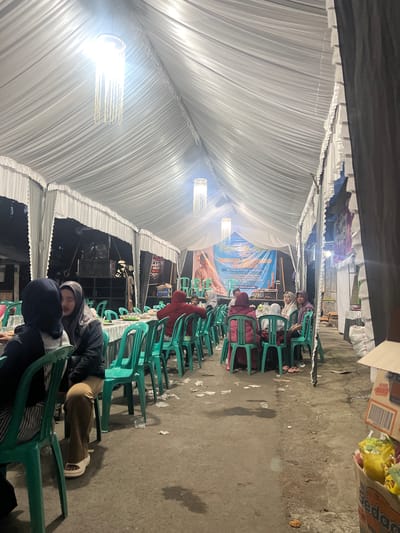 The image depicts an outdoor event tent set up in Wedarijaksa, Indonesia. Several people are seated at tables, some of whom appear to be dining or conversing. The tent is decorated with white fabric draped from the ceiling, and hanging light fixtures illuminate the space. Green plastic chairs surround the tables, and a banner with text in Indonesian is visible in the background. The ground is concrete, and there is some litter scattered around. To the right, a cardboard box filled with yellow plastic bags is present. The overall atmosphere suggests a community gathering or celebration.