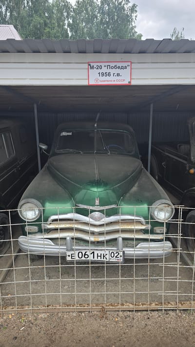 In the city of Bol'shoi Mashlyak, Russia, a vintage green GAZ M-20 Pobeda from 1956 is parked inside a covered, open-air garage. The car is covered in dust, and a white sign with red lettering above it reads "M-20 "Pobeda" 1956 G.V. Made in USSR". The license plate reads "E 061HK 02 RUS". The car is behind a metal fence, and other vehicles are visible on either side. The ground is covered in dirt and gravel.