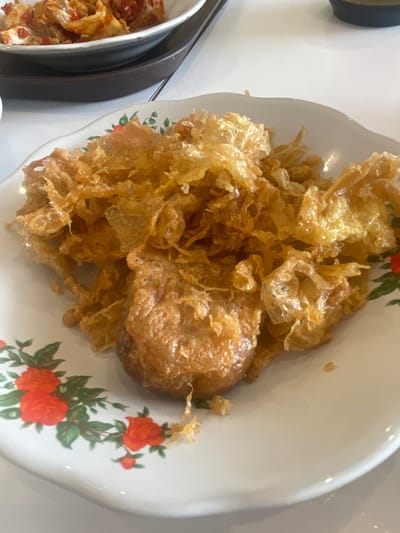 A close-up, eye-level shot displays a white ceramic plate filled with a generous portion of crispy, deep-fried food. The food is golden brown and appears to be a fluffy, delicate batter, possibly resembling crispy fried chicken or a type of fried omelet. A larger, more solid piece of food sits in the center of the plate, also golden brown and fried. The plate itself is adorned with a floral pattern featuring red roses and green leaves along its wavy rim. In the background, a white tabletop is visible, along with a glimpse of another dish containing what seems to be kimchi, also in a white bowl. The setting appears to be a restaurant in Medan, Indonesia, based on the provided contextual information. The lighting is bright, suggesting an indoor environment with overhead illumination. There are no people visible in the image.