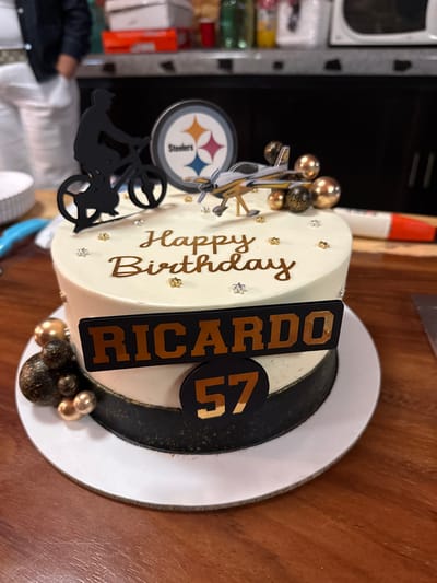 The image shows a decorated cake on a wooden surface. The cake is round and frosted in a light cream color. "Happy Birthday" is written in gold script on the side, along with scattered gold and silver star decorations. A black rectangular plaque with the name "RICARDO" in gold block letters is placed on top of the cake, along with a black circle displaying the number "57" in gold. The cake is also decorated with gold and black spheres. To the left of the name are decorations related to the Pittsburgh Steelers (NFL team), including a Steelers logo and a silhouette of a biker. The cake sits on a white cake plate. The background is blurred but includes glimpses of a kitchen area with cabinets, containers, and an open area. There is no evidence to suggest this photo was taken in San Francisco Lachigoló, Mexico.