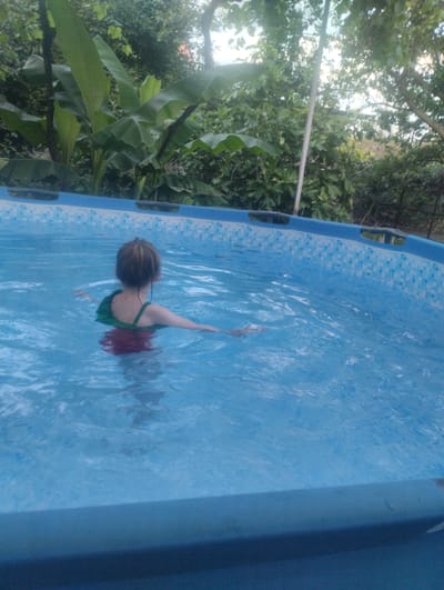 A child with dark hair swims in a blue pool. The child is wearing a green and red swimsuit. Lush green foliage surrounds the pool.