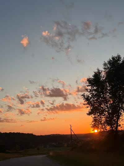This image depicts a rural landscape at sunset, with the sun partially obscured by a silhouetted tree and a utility pole. The sky transitions from a pale blue at the top to a vibrant orange near the horizon, dotted with scattered clouds catching the last rays of sunlight. In the foreground, a paved road curves to the left. Beyond the road, a dark, wooded hillside overlooks a valley containing a small settlement with visible houses and structures. A utility pole with electrical wires stands prominently in the mid-ground, just to the left of the silhouetted tree. The overall scene evokes a tranquil evening in the Ukrainian countryside.