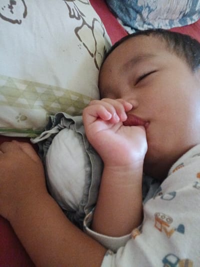 The image shows a close-up of a young child sleeping, with their hand in their mouth. The child is lying on a bed with a patterned pillow and a blanket. The environment appears to be indoors. The child is wearing a white onesie with a graphic print. The overall impression is one of peaceful sleep. This image was taken in Cibungbulang, Indonesia.