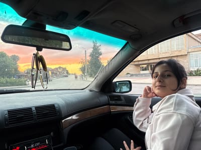 A young woman with dark hair is seated in the passenger seat of a car, resting her chin on her hand and looking at the camera. She is wearing a light pink sweatshirt. The interior of the car is visible, including the dashboard with air vents and a car stereo. Hanging from the rearview mirror are a pair of sunglasses and two air fresheners. Outside the car, through the windshield, a street scene is visible during sunset, with buildings, trees, streetlights, and a few people walking. The sky is filled with vibrant orange and pink hues. The scene appears to be in the city of Letnitsa, Bulgaria.