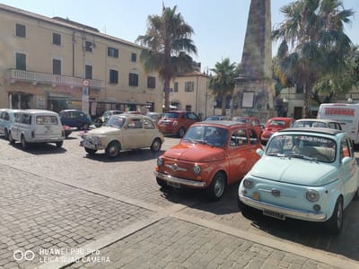 A light blue Fiat 500, an orange Fiat 500, and a beige Fiat 500 are parked in a cobblestone square in Follonica, Italy, near a stone monument. The cars are vintage models. In the background, there are buildings, palm trees, and a white van. The sky is clear and blue. The scene appears to be a sunny day, possibly part of a car show or gathering.