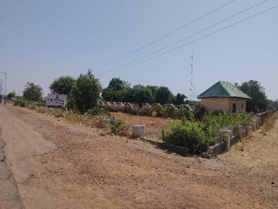 The image shows a dirt road in front of a low-lying building with a green roof and a sign that reads "K.W. ENTERPRISES, PROPS." The building is surrounded by a low wall and a fence. There is a tall antenna in the background. The image is taken at the entrance to the K.W. Enterprises property in Sangaro, Nigeria. The image is taken from a distance, looking down the road towards the building and the surrounding area. The location of the image is approximately latitude 10.834 and longitude 11.429. The image captures the scene on a clear, sunny day, with a few clouds visible in the sky. The road is dusty, and there is some vegetation on the side of the road. There are no people visible in the image. The scene is relatively deserted. The light is bright, and the image is well-exposed.