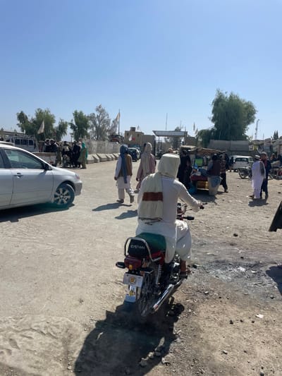 This image depicts a dusty, sunlit outdoor scene in Spin Buldak, Afghanistan, likely at a border crossing or checkpoint area. In the foreground, a man wearing white traditional attire and a white head covering with a brown and white striped shawl is seated on a red "CROWN" brand motorcycle, viewed from the rear. The motorcycle has a visible license plate and chrome exhaust. The ground is unpaved, arid, and marked with tire tracks and debris, with long shadows cast by people and objects indicating bright overhead sunlight.
In the mid-ground, a silver or white sedan is partially visible on the left. Numerous pedestrians, dressed in traditional Afghan clothing, are walking or gathered across the scene. A cluster of individuals is observed interacting near the left mid-ground. Further right, what appears to be a market stall or vendor cart is present, with more people and another motorcycle in the vicinity.
The background features structures consistent with a border facility: concrete barriers, an archway-like entrance, and various vehicles including trucks and cars. Multiple light-colored flags, possibly white with a yellow emblem, are visible on poles and structures throughout the background. Sparse trees with light green foliage are also present. The sky is clear and blue.