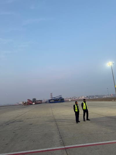Two airport workers in high-visibility vests stand beside a tarmac. Behind them, a blue cargo loader with the word "Indigo" is visible. Other airport vehicles and infrastructure are blurred in the background due to fog or haze. A tall light pole is on the upper left. The sky is light blue with a gradient of grey near the horizon. The tarmac is concrete with red lane markings.