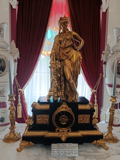 The image captures an ornate gilded clock on a black pedestal, featuring a central dial and intricate gold detailing. To the left and right of the clock are tall, decorative golden candelabra, each holding a tall, slender gray candle. Behind the clock, a draped red curtain with gold trim frames a brightly lit window with stained-glass elements in hues of red, blue, and yellow. Flanking the clock and candelabra are large, white porcelain vases adorned with intricate gold patterns and dark red accents. At the bottom of the image, a plaque with Russian text is visible. The overall setting appears to be an interior with a marble-topped surface. This clock is located in the museum exhibition of Kosava, Belarus, part of the collection of the Kosava Palace.
