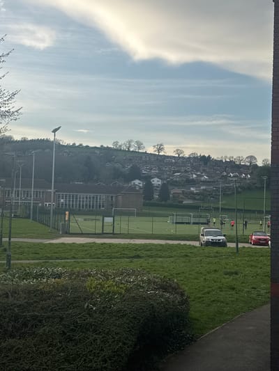 The image shows a view from a window, likely in Welshpool, United Kingdom, as indicated by the presence of football/sports pitches in the background. The sky is predominantly blue with wispy clouds. In the middle ground, there is a cluster of buildings, possibly houses, on a slight hill. Further in the background, sports pitches are visible, with people appearing to play or engage in activities. Vehicles, including a red van or car and a white van, are parked alongside the pitches. The immediate foreground is occupied by a hedge or bushes. The top and bottom of the image are framed by a brick wall or other building structure, suggesting an indoor viewpoint.