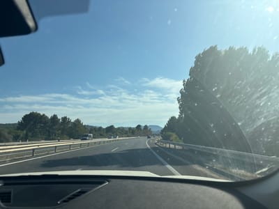 The image captures a view from inside a vehicle traveling on a highway. The perspective is from the driver's seat, looking forward through the windshield. The highway has multiple lanes, divided by white lines, and is bordered by a metal guardrail. On the left side of the highway, there is a median with vegetation and trees, and further back, hills covered in trees. On the right side, dense trees line the road. The sky is a clear blue with some scattered clouds. Several vehicles are visible on the highway, including cars and a van. The sun is shining brightly, creating glare and lens flares on the windshield, obscuring some details. The foreground shows the dashboard of the vehicle. The scene is consistent with a highway setting outside of a populated area.