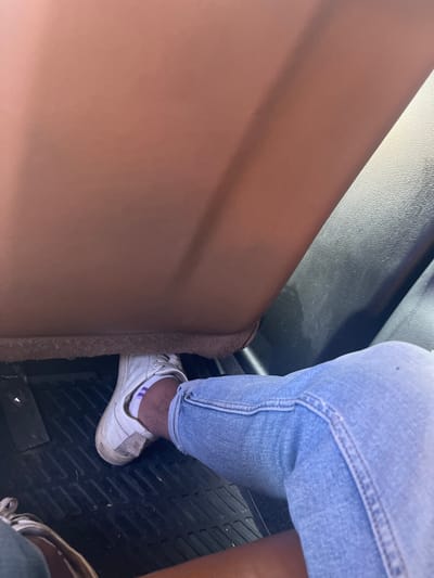 This image shows a person's legs and feet inside a vehicle. The person is wearing blue jeans and white sneakers with purple stripes on the socks. Their right foot is resting on what appears to be a car's floor mat, with a portion of the car's interior visible, including a brown seat and black dashboard. There is no visible indication of the city of Yasen, Bulgaria, within the image.
