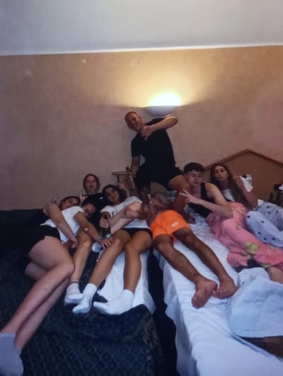 The scene depicts seven young adult females and one young adult male gathered in a room in Aktash, Uzbekistan. The individuals are positioned on or around beds covered with white and dark patterned bedding. A male, centrally located in the upper portion of the frame, stands on a dark patterned bed, wearing a black t-shirt and black shorts, holding a dark bottle in his left hand and making a 'thumbs up' gesture with his right. To his right, another male in orange shorts lies barefoot on a white-sheeted bed. Five females recline on the beds surrounding them. One female on the left, wearing a white t-shirt and dark shorts, holds a green bottle. Another female on the far right wears pink patterned pajama bottoms. Clothing includes casual wear, such as t-shirts and shorts; white socks are visible on two females, and at least two individuals are barefoot. The room features light-colored walls, with a wall-mounted sconce providing warm upward illumination. A dark wooden headboard or furniture piece is partially visible on the right.