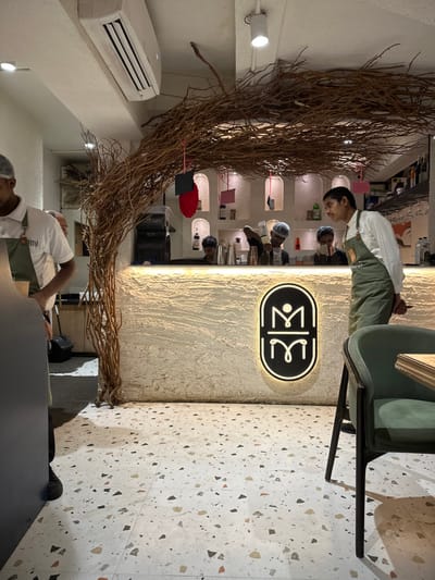The image captures the interior of a contemporary cafe or bar establishment. While the city is Kalyan-Dombivli, India, the exact location within the city is not discernible from the provided visual information.
The scene focuses on a prominent bar area. The bar counter features a light-colored, textured surface with a backlit, black oval-shaped sign displaying a white stylized "M" logo centrally. Overhead, a decorative structure made of intertwined dry tree branches forms an archway, with small red and black tags suspended from it. Behind the bar, a white wall contains multiple arched recesses housing bottles and bar equipment.
The floor is covered with white terrazzo-style tiles featuring multi-colored flecks of black, gray, orange, and beige. The ceiling is white, fitted with an exposed air conditioning unit, recessed spotlights, and a cylindrical track light.
Five male individuals, presumed to be staff members, are present. In the left foreground, a man wearing a white polo shirt, a dark green apron with "Zimi" embroidered on it, and a hair net is partially visible, facing away. Behind the bar, a staff member wearing a hair net and a dark apron is actively pouring liquid from a white squeeze bottle into a metal shaker. Two other staff members, also in hair nets, are partially visible further right behind the bar, engaged in preparation. To the right of the bar, in the mid-ground, a fifth staff member wearing a white long-sleeved shirt and a dark green apron stands upright, facing right with his hands clasped behind his back.
In the right foreground, a dark green upholstered armchair with dark wooden legs is partially visible, indicating customer seating. Various bottles, shakers, and glassware are visible behind and on the bar counter.