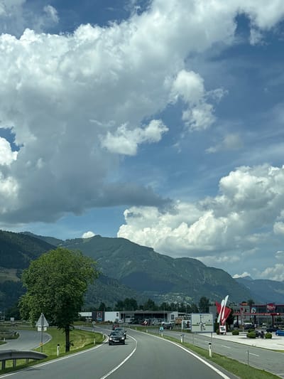 The image depicts a view from a car driving on a road in Bruck an der Großglocknerstraße, Austria. In the foreground, a black car is driving on a paved road with white lane markings. To the left, a large, leafy green tree stands beside the road, with a traffic sign in front of it. Further down the road, several buildings are visible, including a large supermarket with the "ALDI" logo and other commercial establishments. A prominent red and white sculpture, resembling cutlery, stands in front of one of the buildings, labeled "maxi restaurant". Behind these buildings, a range of lush green mountains rises, covered with trees, and a blue sky with scattered white clouds fills the upper portion of the image. The scene suggests a sunny day, with the mountains serving as a backdrop to the urban development.