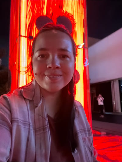 In the city of Koto, Japan, a woman is photographed in a selfie outdoors at night, illuminated by vibrant red and yellow lights. She is wearing a plaid shirt and mouse ears, smiling at the camera. The background features a tall, illuminated structure with vertical red and yellow lights, and a person standing in the distance. The ground is also lit with a bright red glow, suggesting an artificial environment.