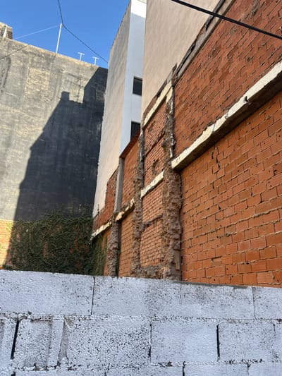 This image captures an outdoor scene with a focus on architectural elements and construction. The foreground features a wall constructed from large, white, textured blocks, with visible lines indicating individual units. Adjacent to this is a section of exposed red brickwork, which appears to be part of a wall or ceiling. The bricks are laid in a traditional pattern, and some of the mortar is crumbling, suggesting ongoing construction or disrepair.
The background reveals more of the surrounding environment. A light-colored building with windows is visible on the left, partially obscured by a dark, textured wall. This dark wall has a rough surface and shows signs of weathering. Sunlight casts a strong shadow across this wall, indicating it is likely daytime. Further to the left, a clear blue sky is visible, along with electrical wires and two white vertical structures that resemble antenna mounts.
A significant portion of the image is dominated by the red brickwork, which is angled upwards, giving a sense of looking up or into a recessed space. There are patches of plant growth, specifically a dense vine, clinging to the lower sections of the brick wall on the right side. The overall impression is one of an urban or semi-urban setting undergoing some form of development or renovation. The lighting suggests a sunny day, possibly late morning or early afternoon, given the distinct shadows. No people or explicit text are visible in the image. The location context provided is Castelló De La Plana, Spain.