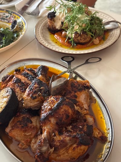 A top-down shot reveals two plates and a small bowl of food. On the left, a plate holds fried items topped with white sauce and herbs. To the right, a silver platter is laden with roasted chicken and vegetables in a rich, oily sauce. A pair of silver tongs and a spoon rest atop the platter. The bottom left corner shows a glimpse of another dish, filled with green herbs and possibly lemon. The arrangement suggests a shared meal.