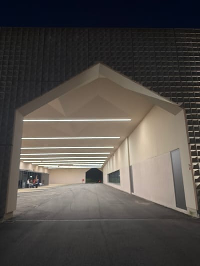 The image displays a modern architectural structure at night in Penafiel, Portugal. The central feature is a large, gabled archway, functioning as an illuminated covered passage or entrance. The building's exterior surfaces, above and flanking the archway, are clad in dark, possibly charcoal-grey, square-patterned relief tiles. The interior of the archway features light-toned, possibly off-white, faceted walls and ceiling. Nine parallel horizontal LED strip lights are embedded in the ceiling, providing consistent illumination along the passage.
On the left side of the archway, a glass-fronted section reveals a group of approximately four to five individuals, some seated, within what appears to be an interior waiting area. The ground beneath the archway consists of dark asphalt pavement, exhibiting subtle tire marks. A linear drain grate is visible across the pavement in the immediate foreground. The interior right wall of the passage is light-toned, featuring a long, horizontal window providing a view into an adjacent space, and further along, a dark grey vertical door. The passage extends into the distance, terminating in another dark, gabled opening, within which a single red light source is discernible.
