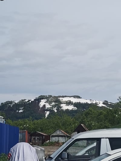 The image depicts a cloudy day with a view of a snow-capped mountain in the distance. In the foreground, there are several structures, including a blue corrugated metal fence, some small sheds or garages, and a white van. A grey tarp covers an object in the lower left corner, with text visible that appears to say "TIC CAT". Trees and greenery are visible between the foreground structures and the mountain. The scene appears to be an outdoor area with some industrial or storage elements, located in or near Termal'nyy, Russia, as indicated by the presence of the snow-capped mountain, a common feature of the volcanic landscapes in that region.