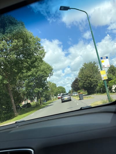 The image is taken from the perspective of someone inside a vehicle, looking out through the windshield. The scene depicts a sunny day in Maarssen, Netherlands, with a clear blue sky dotted with fluffy white clouds. A road stretches into the distance, lined with lush green trees on both sides.
In the foreground, the dashboard of the car is visible. On the right side of the road, a modern streetlight with a curved arm stands tall. Attached to the streetlight pole are two yellow signs, one at the top depicting a bus icon and text, and another below with more text and a bus icon. A grey garbage bin is situated on the grass verge next to the road, near the signs.
Further down the road, several cars are visible. A silver hatchback is in the immediate lane ahead, followed by a white van and other vehicles further in the distance. On the left side of the road, houses and fences are partially obscured by trees. The overall impression is a suburban or semi-rural environment with active road traffic.
