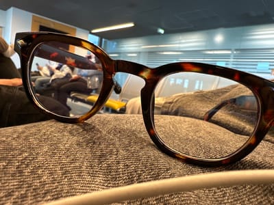 A pair of tortoise-shell patterned eyeglasses rests on a textured, grey fabric surface. The image is captured from a low perspective, looking through and past the frames into a bright, modern indoor environment.
Through the lenses and in the background, a light-colored, reflective floor shows a prominent horizontal streak of light. A light-toned railing or barrier stretches across the mid-ground. Beyond this, blurry reflections of several people are visible, suggesting a public area with activity. Distant, indistinct signage and an orange-brown object, possibly art or another sign, can also be perceived.
A notable detail on the eyeglasses is a small, metallic, pyramidal stud located on the outer corner of the right frame. The brand name "New York Montage" is clearly visible and legible on the inside of the right temple arm of the glasses. The contemporary architecture, polished surfaces, and reflections of individuals in the background, set within Weeze, Germany, indicate a bustling public concourse or waiting area, likely an airport terminal. The scene is well-lit, suggesting daytime.