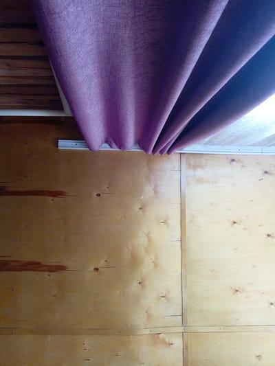 The image shows an interior scene with wood paneling on the walls and a purple curtain hanging from a track near the ceiling. The wood paneling appears to be plywood with visible knots and some stains. The curtain is draped and folded, creating shadows and highlights. The overall impression is of a rustic or unfinished interior space. The location in Churapcha, Russia, suggests a cold climate, and the interior might be part of a dwelling or a structure designed for warmth.