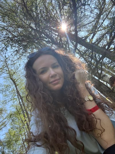 A low-angle, medium shot captures a woman with long, curly brown hair and sunglasses perched on her head, looking up towards the sun filtered through the branches of a larch tree. She is wearing a light-colored t-shirt and a red wristband with Cyrillic text that appears to read "Хорошо Быть" (It's Good to Be). A gold watch is on her left wrist. The background consists of a bright blue sky and numerous branches and needles of the larch tree. Another person, possibly a child, is partially visible in the upper right background, sitting in the tree. The scene appears to be outdoors, in a wooded area, potentially at the Bichiktu-Boom location in Russia, given the context.
