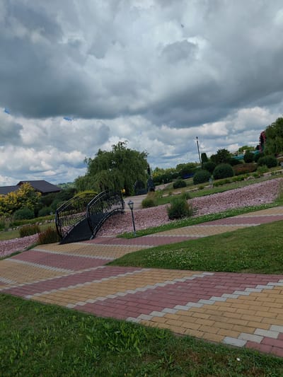 The image depicts an outdoor park scene under a cloudy sky. In the foreground, a pathway paved with red, yellow, and gray interlocking stones winds through lush green grass. To the left and center, a black ornamental metal bridge with intricate scrollwork spans across a dry streambed filled with light pink stones. A lamppost stands near the bridge. In the background, a large weeping willow tree dominates the landscape, with a building featuring a dark brown roof visible behind it. Further in the distance, more trees, bushes, and decorative plants are arranged on a tiered hillside. The scene appears to be a well-maintained public space, likely in the city of Kovalivka, Ukraine.