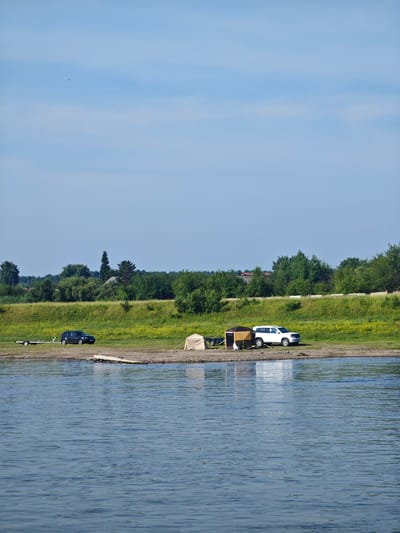The image depicts a serene riverside scene on a clear day. In the foreground, a wide expanse of blue water with gentle ripples occupies the bottom half of the frame. The opposite bank features a gently sloping grassy hill covered with scattered yellow wildflowers, leading up to a dense line of green trees. Beyond the trees, glimpses of buildings with red roofs suggest a settlement.
On the sandy bank in the middle ground, two SUVs are parked. A dark blue SUV is positioned on the left, attached to a trailer. To its right, a light-colored SUV is parked near a small campsite setup. This campsite includes a beige tent and a brown and tan canopy structure. The ground is a mix of sand and pebbles. The sky above is a bright, clear blue with a few wispy clouds and a tiny dark silhouette of a bird in flight. The overall impression is one of a quiet outdoor recreational area, likely near the village of Galanino, Russia, given the context. There are no people visible in the image.