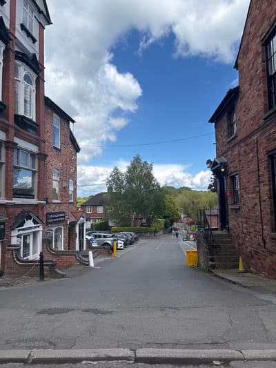 The image captures a street scene in Knutsford, United Kingdom. The street is flanked by brick buildings with storefronts, including one for Clare & Illingworth, a contact lens practitioner. Cars are parked along the side of the road. The sky is visible between the buildings, showing a mix of blue sky and white clouds. In the distance, greenery suggests a park or wooded area. People can be seen walking along the street. Street furniture includes yellow traffic cones and bollards. The road surface shows signs of wear and patching. The scene is captured at eye level and the photograph is shot at a vertical angle.