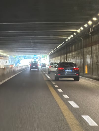 This image depicts the interior of a highway tunnel with traffic. The perspective is from a vehicle moving forward within the tunnel. Several cars are visible, including a dark SUV with a license plate reading "CR 241CK" in the foreground, a white SUV behind it, and other vehicles further ahead. The tunnel is illuminated by overhead lights, and the road has clear lane markings, including a solid yellow line and dashed white lines. The tunnel walls appear to be made of concrete or stone. In the distance, the tunnel opens to daylight, revealing trees and structures outside. The location is specified as the city of Cardano al Campo, Italy.