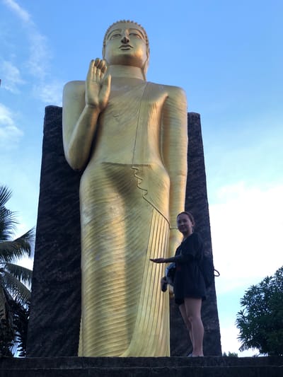 A low-angle shot presents a golden, reclining Buddha statue set against a blue sky with wispy clouds, suggesting daylight. The statue is resting on a dark, textured platform. A person stands at the statue's side, leaning slightly towards the camera with a small backpack over their shoulder. Palm trees are visible at the bottom and lush foliage at the top right of the frame. This statue is located in the city of Dondra, Sri Lanka.